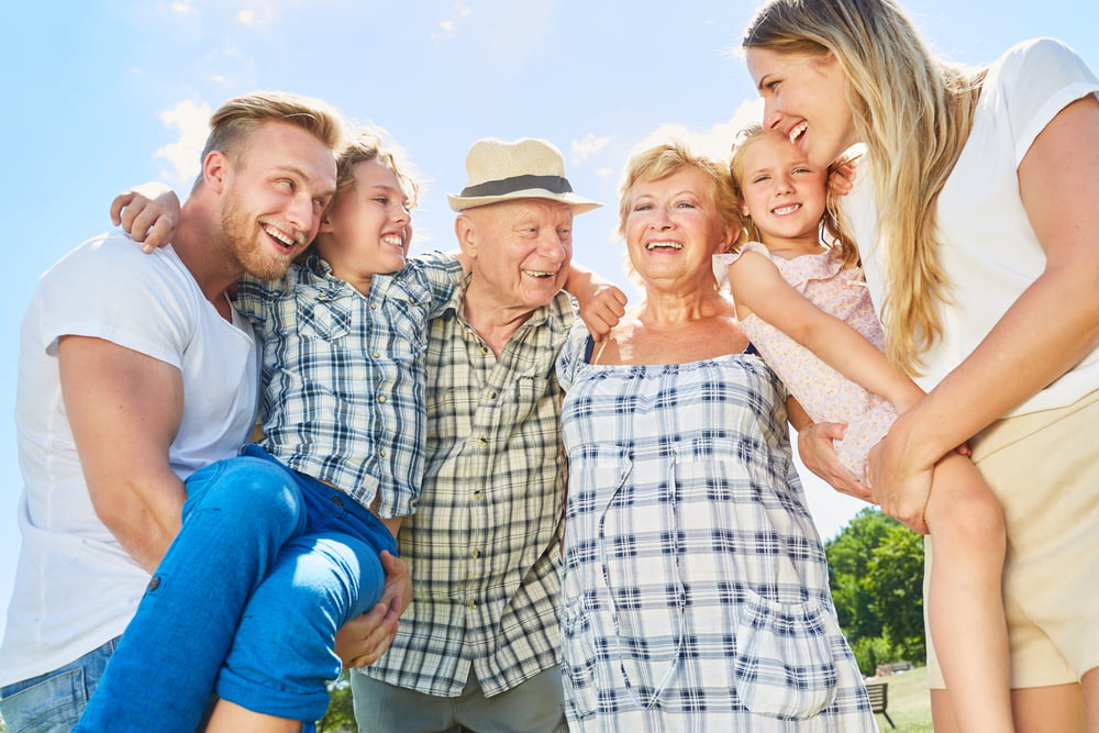 A happy multi-generational family stands together outdoors, smiling and embracing under a bright blue sky - Primary Care Sheridan WY A happy multi-generational family stands together outdoors, smiling and embracing under a bright blue sky - Primary Care Sheridan WY